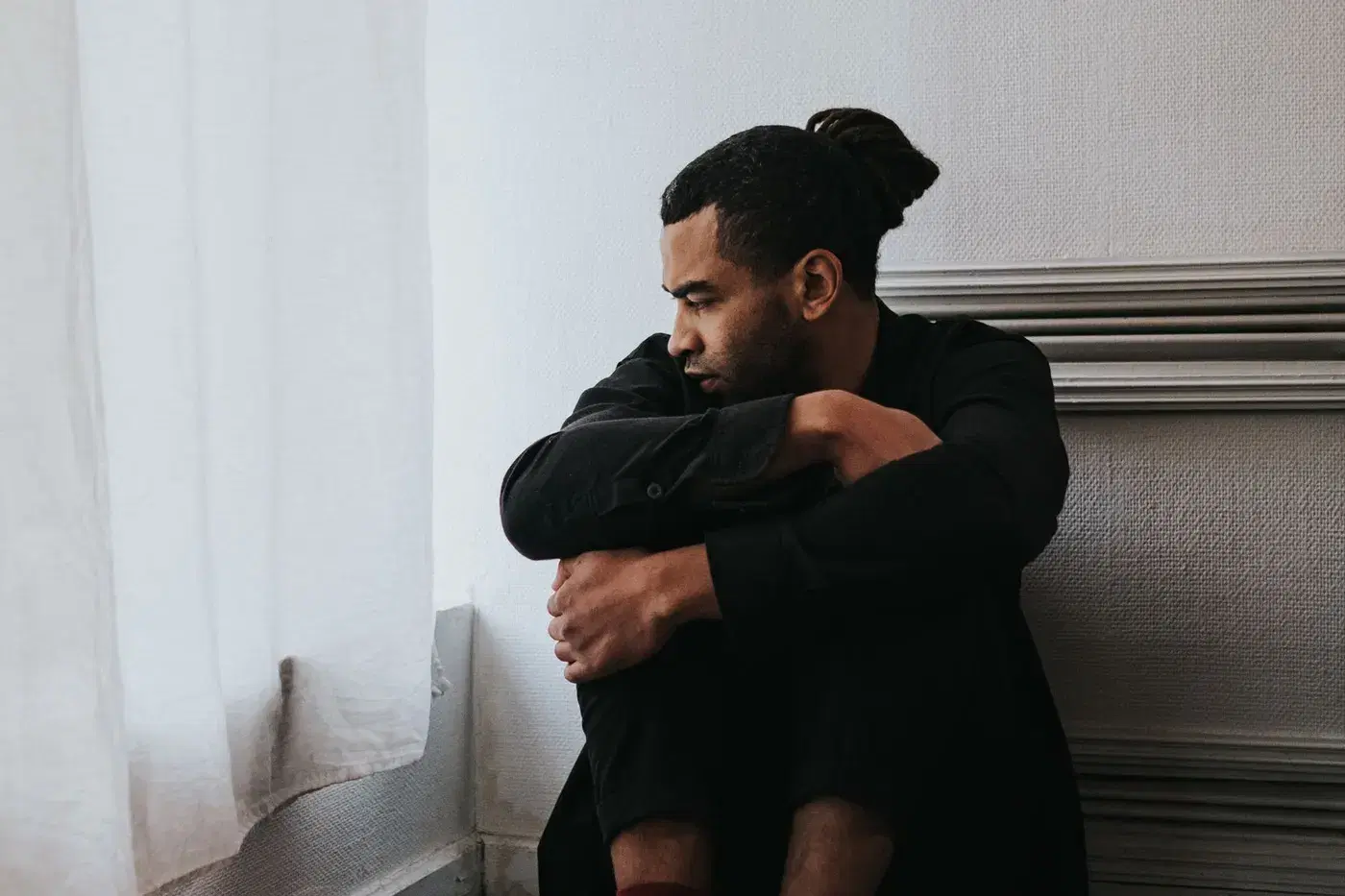 A man sitting alone by a bright window, arms folded, looking inward