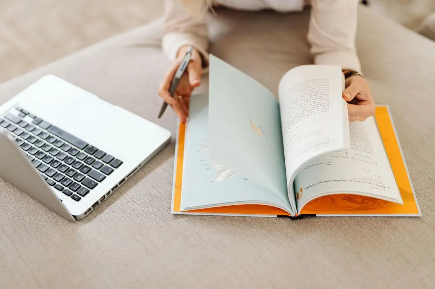 An open laptop beside an open book, with hands marking a page